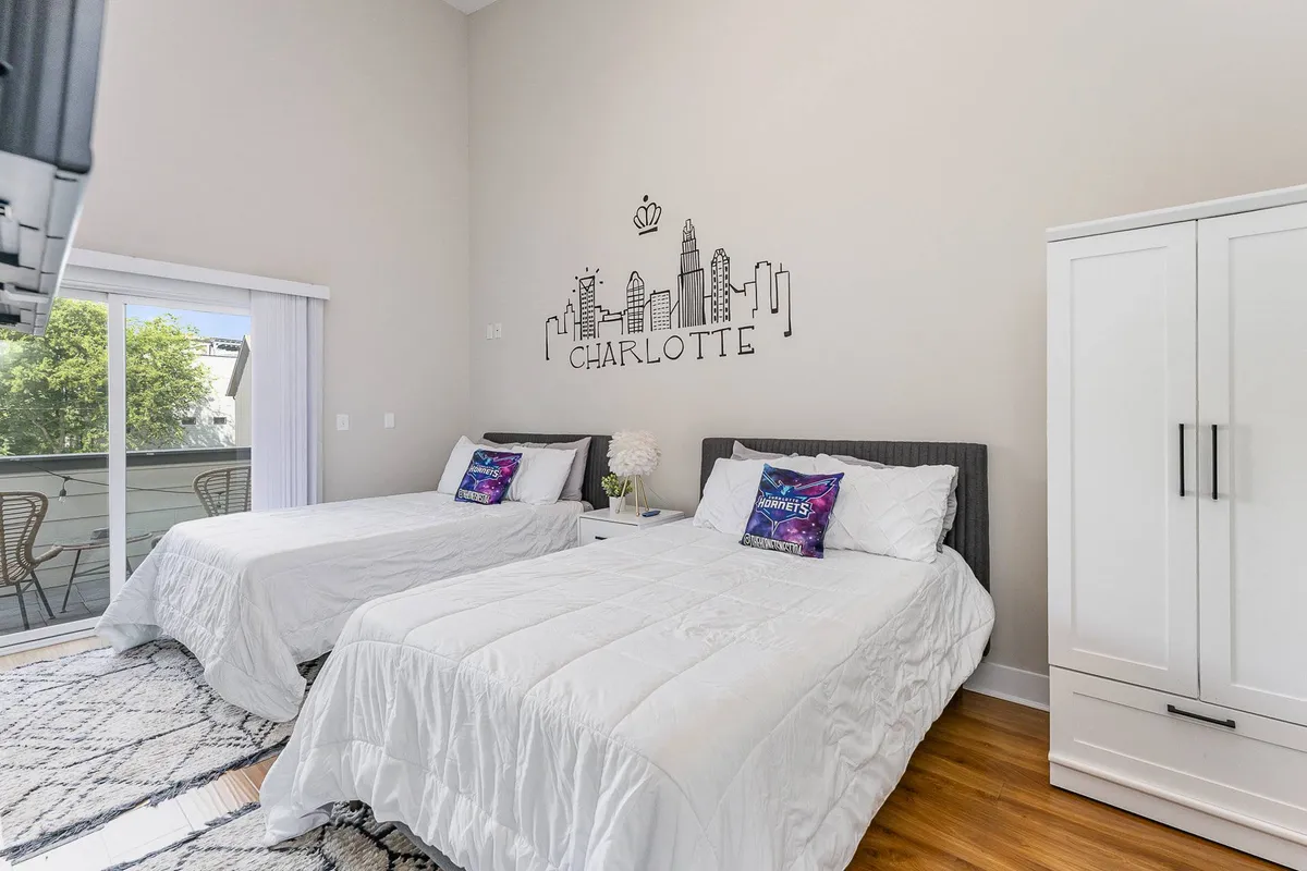 A bright and inviting bedroom featuring two beds with colorful decorative pillows, a bold city skyline mural above the headboards, and a wardrobe. Soft morning light filters in from a private balcony, visible through glass doors, offering a glimpse of the city skyline beyond.