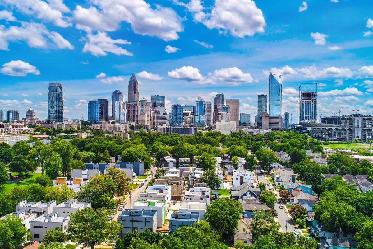 A sweeping view of Charlotte's city skyline framed by green parks and vibrant residential neighborhoods. The scene is energetic, showcasing the blend of urban excitement and natural spaces. People can be seen enjoying outdoor activities like picnicking and exploring local cafes, reflecting a lively community atmosphere.