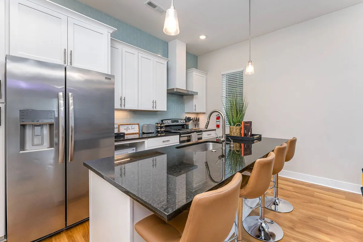 A sleek kitchen scene with a stainless steel refrigerator, black granite countertops, and bar stools lined up at the breakfast bar. The room exudes a contemporary, welcoming vibe suited for entertaining, casual dining, or prepping snacks before a night out.