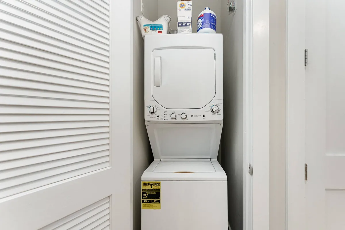 The photo displays a stacked washer and dryer unit with laundry supplies neatly organized nearby, set in a bright, functional laundry area.