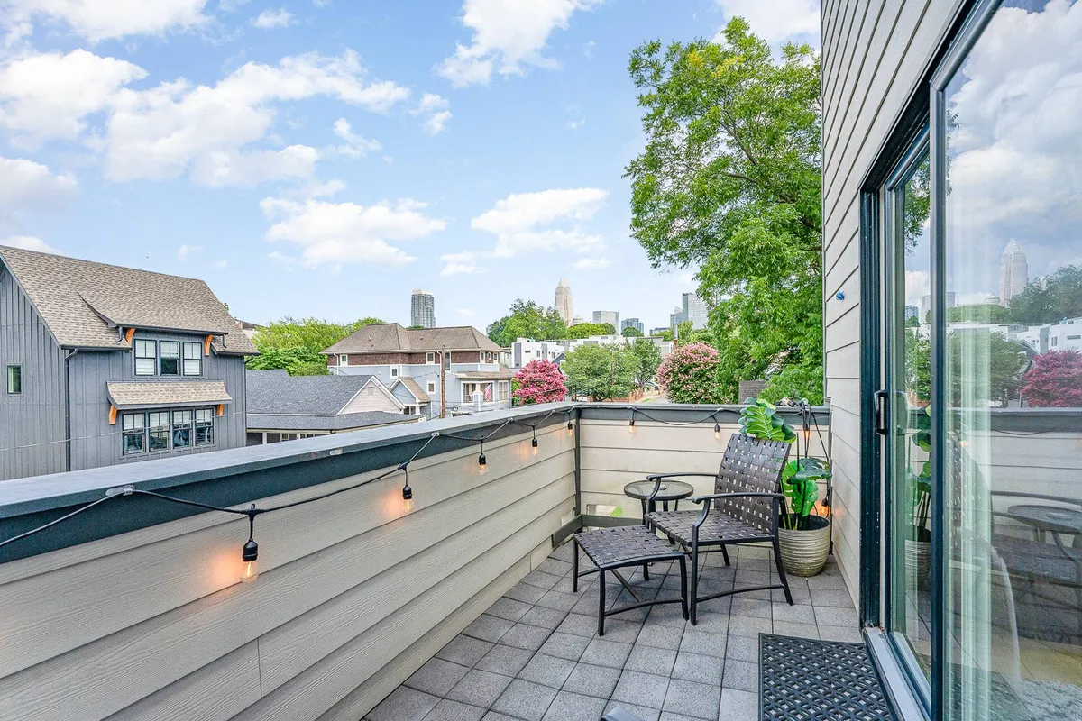 The image showcases the exterior of a stylish single-family home in Uptown Charlotte. The hero shot features a spacious balcony adorned with cozy seating and glowing string lights, offering panoramic views of the city skyline framed by lush greenery. The setting radiates a serene, inviting atmosphere—perfect for relaxing, entertaining, or simply soaking in the urban charm as the city lights twinkle after dark.