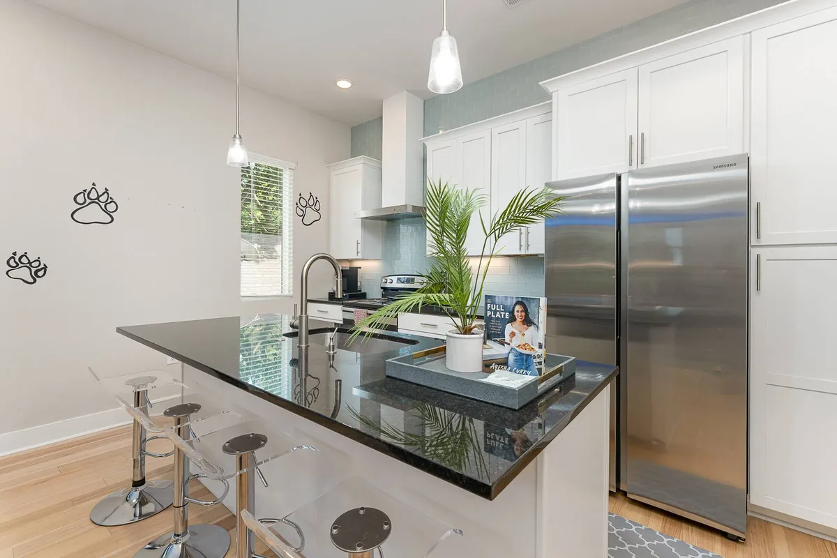A cheerful kitchen scene showing a kitchen island with comfortable seating, stainless steel appliances, a decorative plant, and playful paw print wall art. The open layout hints at easy movement between kitchen, dining, and living areas, ideal for entertaining or family meals.