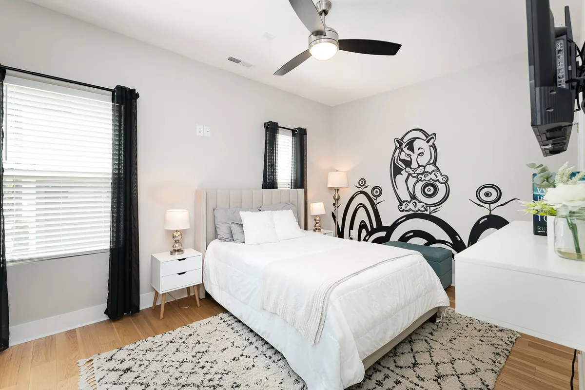 A stylish bedroom with light bedding, a large graphic mural on the wall, decorative side tables with lamps, and a TV mounted opposite the bed. Sunlight fills the space, highlighting the unique, playful design elements and creating a cozy, personal ambiance.
