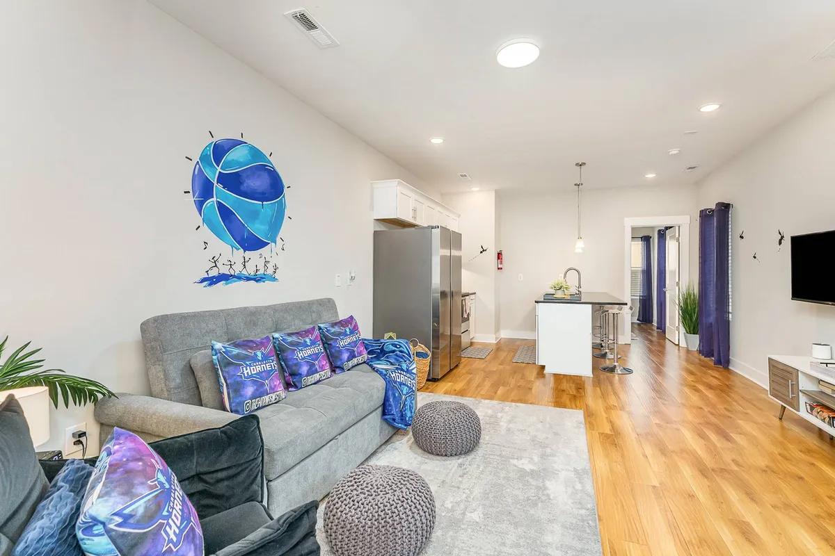 A vibrant living area with a striking basketball mural on the wall, a sofa adorned with colorful throw pillows, and a sleek modern kitchen island in view. The space exudes a playful, energetic vibe perfect for group gatherings.