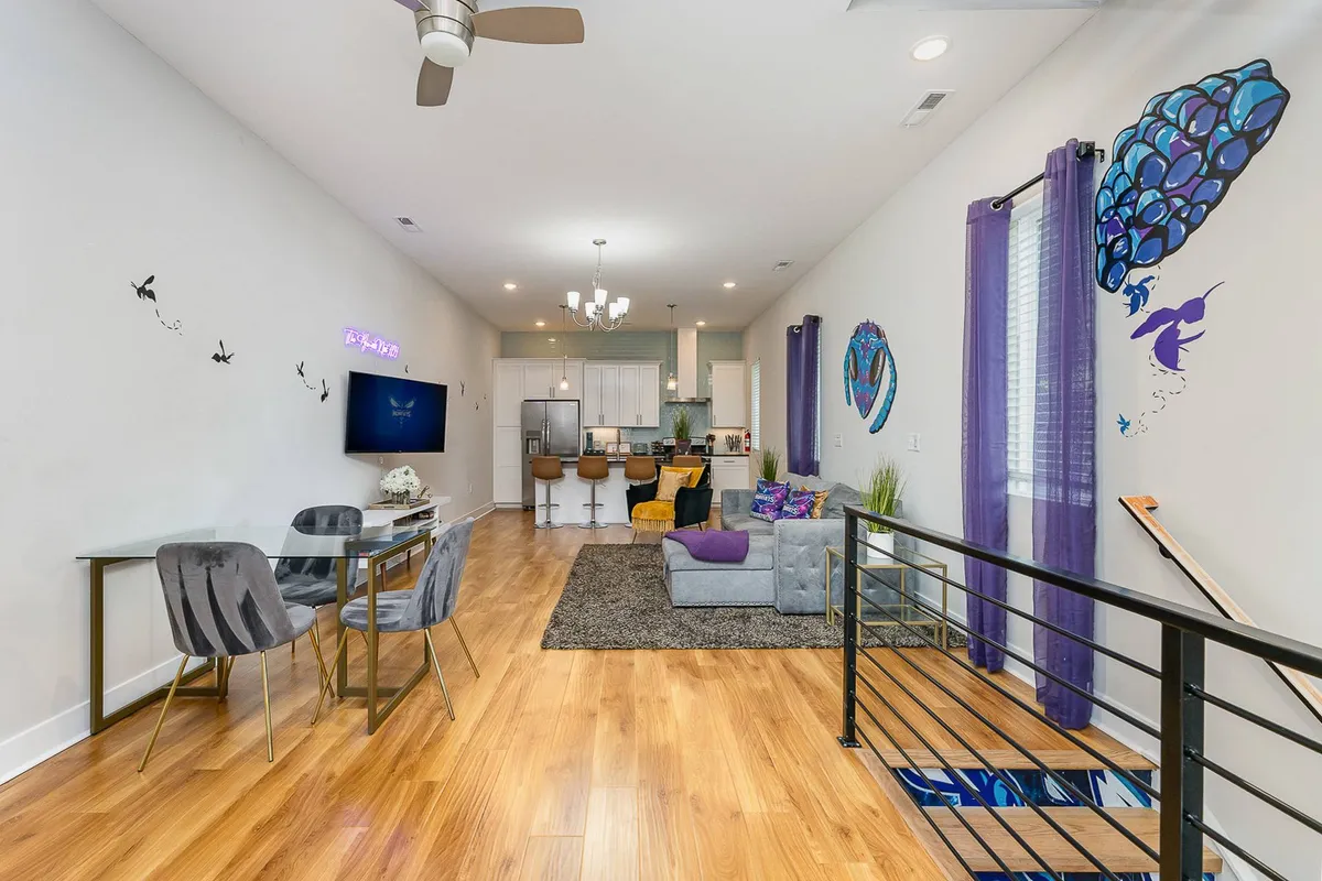 A lively open-plan living and dining area featuring colorful wall art, modern furniture, a spacious dining area with bar stools, and abundant natural light. The setting looks ready for social gatherings, game nights, and movie marathons.