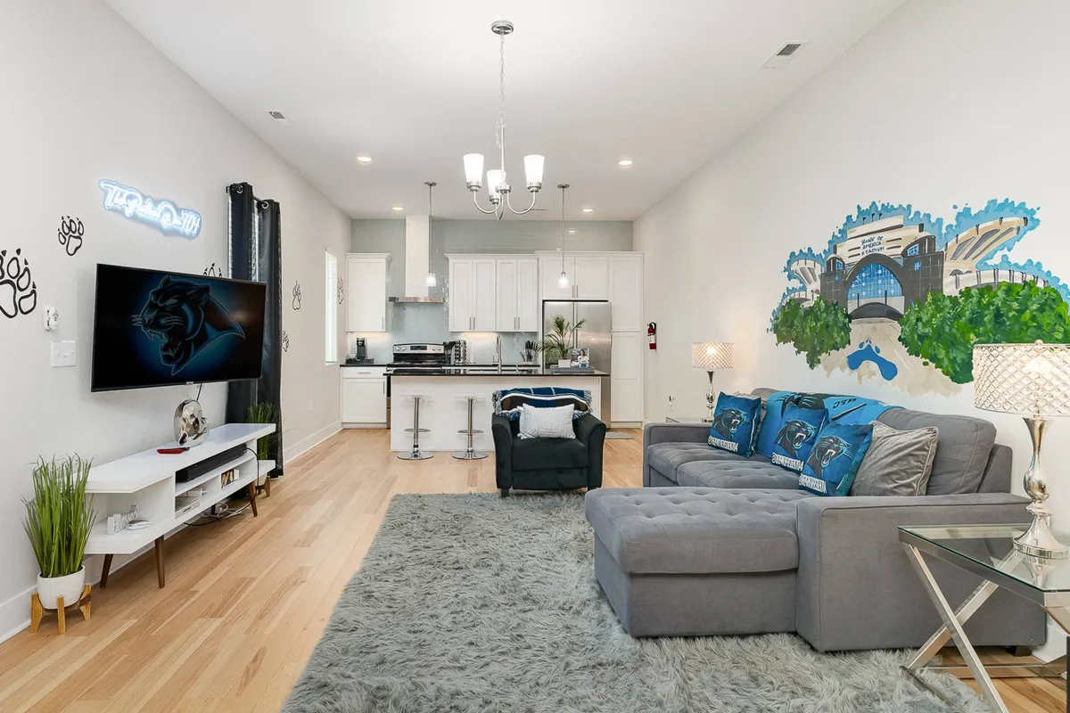 A cozy, contemporary living space with a sofa and decorative pillows, flat-screen TV, kitchen island, and a striking wall mural. There’s a clear division between living and cooking zones, inviting guests to both relax and entertain.