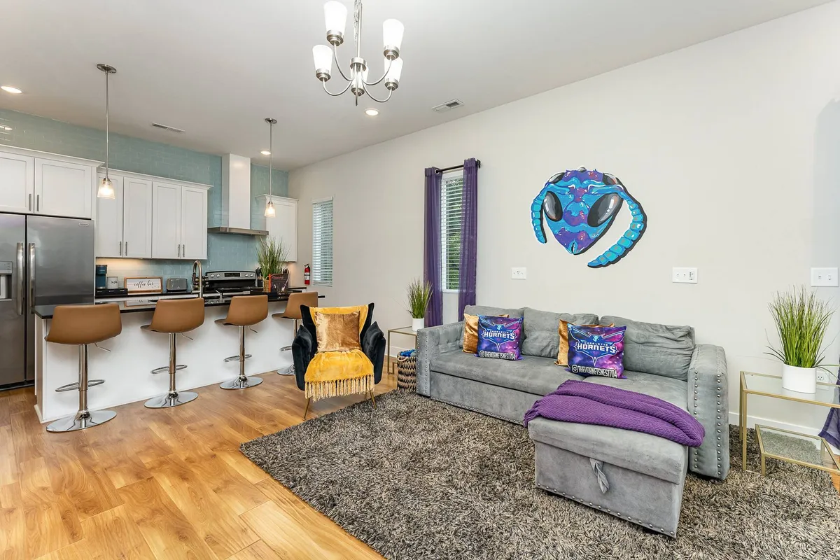 A modern open-plan living and kitchen area featuring a stylish sofa, contemporary island, statement chandelier, and vibrant artwork. The space is set up for social gatherings and shared meals, inviting guests to relax and celebrate together.